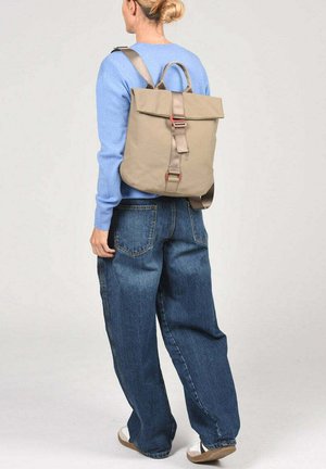 Mujer con un jersey azul claro, vaqueros sueltos azules, zapatillas blancas y una mochila beige con hebilla frontal, de pie frente a un fondo liso.