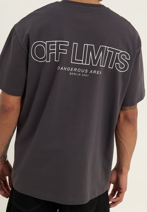Persoon die een donkergrijs T-shirt draagt met de tekst "OFF LIMITS" en "DANGEROUS AREA BERLIN 2001" in wit op de achterkant gedrukt.