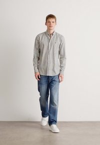 Camicia a quadri verde e bianca, abbinata a jeans blu e sneakers bianche; in piedi davanti a uno sfondo chiaro e semplice.