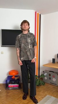 Camisa de manga corta con estampado de camuflaje en verdes apagados, combinada con pantalones de pierna ancha negros; usada con zapatos marrón oscuro. Se ven pocos accesorios.
