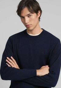 Maglione a maglia blu marina con scollo rotondo, caratterizzato da un logo discreto sul petto. Tessuto morbido e maniche lunghe.
