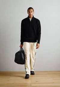 Svart glidelåsgenser, elfenbensfargede joggebukser med sidestriper, svarte sko og en svart treningsbag. Treverk gulv og lys grå bakgrunn.