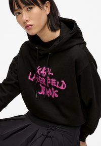 Felpa nera con cappuccio in tessuto morbido, con logo sprayato rosa "KARL LAGERFELD JEANS," abbinata a una gonna plissettata nera.