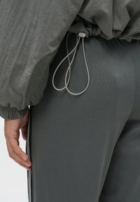 Άτομο ντυμένο με σκούρες γκρι παντελόνες με ελαστική μέση και κορδόνια, και αντίστοιχο γκρι μπουφάν με ελαστικές μανσέτες, το χέρι χαλαρό στο πλάι.