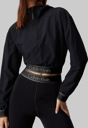 Kobieta ubrana w czarną, krótką bluzę na zamek i legginsy z wysokim stanem, oba zdobione taśmami z logo Calvin Klein.