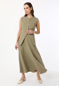 Robe sans manches olive avec un pli subtil, ornée de six boutons dorés sur le devant. Tissu doux et silhouette en A. Chaussures à talons blocs crème.