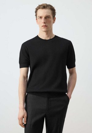 Joven con el pelo rubio corto que lleva un jersey negro acanalado de manga corta y pantalones oscuros, de pie con una mano en el bolsillo.