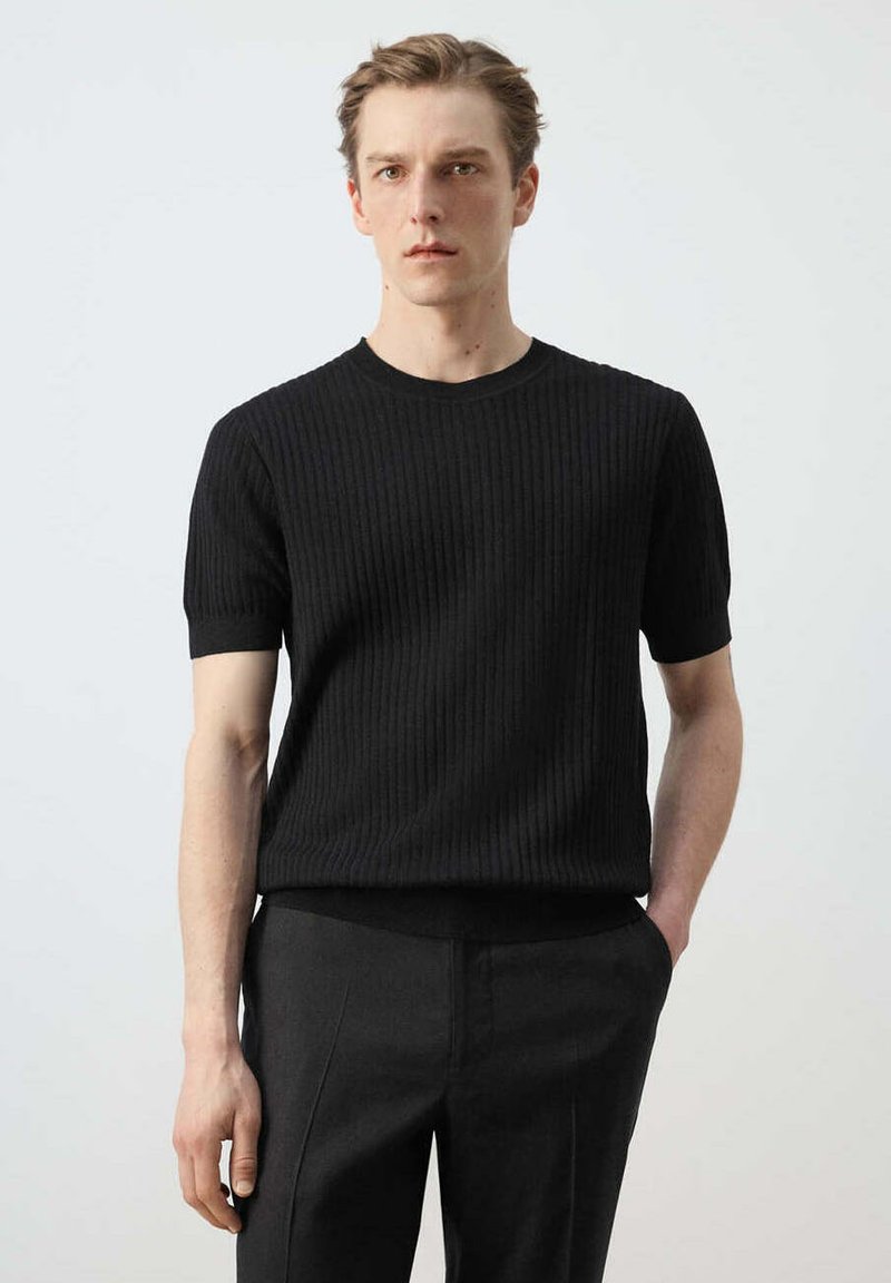 Jeune homme aux cheveux blonds courts portant un pull noir côtelé à manches courtes et un pantalon foncé, debout avec une main dans la poche.