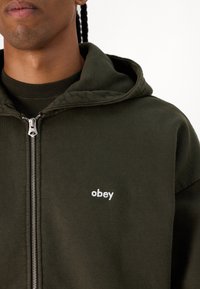 Tamnozeleni dukserica sa zatvaračem, prednjim džepom i bijelim vezom "obey" na prsima. Mekana, teksturirana tkanina, ležeran kroj.