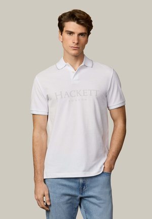 Joven con una camiseta polo blanca de Hackett London y jeans azul claro, de pie contra un fondo beige liso con una mano en el bolsillo.