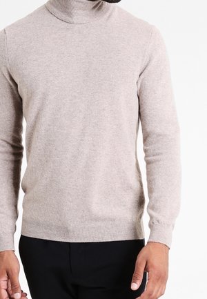 Homme portant un pull à col roulé beige clair et un pantalon noir, se tenant devant un fond blanc uni.