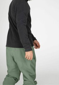 Camicia a maniche lunghe in pile nera abbinata a pantaloni da esterno larghi di colore verde chiaro. Caratteristiche includono un colletto alto e tasche laterali.