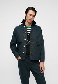 Chaqueta azul marino oscuro con cuello de pana, cierre de botones y dos bolsillos de parche, llevada sobre un suéter a rayas y pantalones a juego.