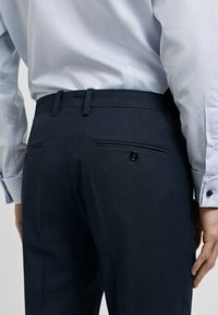 Pantaloni bleumarin cu o textură netedă, cu un design frontal plat, bucle înguste pentru curea și două buzunare la spate cu închidere cu buton.
