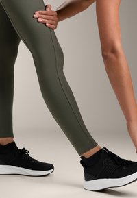 Leggings verde oliva con una superficie liscia e un design aderente, abbinati a scarpe da ginnastica nere con suole e lacci bianchi.