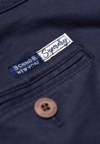 Navy blue chino fabric with a stitched pocket featuring a Superdry label. Includes a brown button with engraved details. Smooth texture.