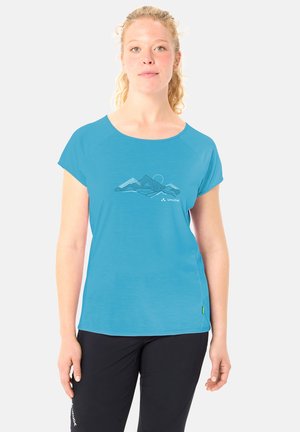 Junge Frau mit blonden, lockigen Haaren, die ein blaues kurzärmeliges Shirt mit Bergdesign und schwarze Hose trägt und vor hellem Hintergrund steht.