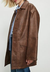 Femme portant un manteau en cuir marron oversize boutonné sur une chemise rayée bleue et un jean bleu, se tenant contre un fond uni.