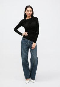 Črna dolga majica z naborki, kombinirana z visokim pasom, ohlapnimi modrimi jeans hlačami in belimi čevlji z ostrim prstom.