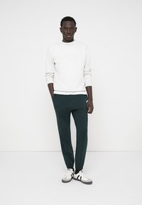 Felpa bianca strutturata con collo rotondo abbinata a pantaloni della tuta tapered di colore verde scuro e scarpe sportive bianche con dettagli neri.