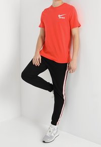 Osoba v červeném tričku Nike "Just Do It", černých kalhotách s růžovými pruhy a bílých teniskách Nike, opřená o bílou zeď.