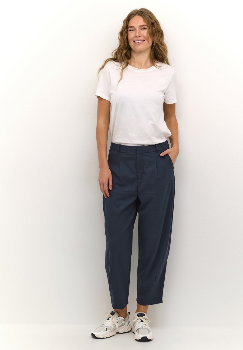 Pantalons navy avec plis à l'avant, poches latérales et jambes fuselées, associés à un t-shirt blanc uni et des baskets de couleur claire.