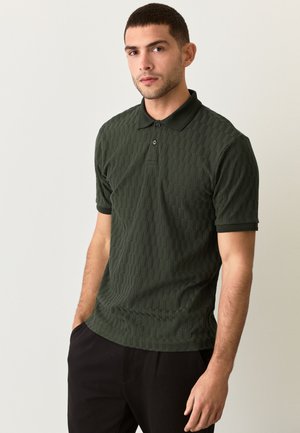 Junger Mann mit kurzen Haaren, der ein dunkelgrünes, strukturiertes Poloshirt und schwarze Hosen trägt, steht mit den Händen in den Taschen vor einem einfarbigen Hintergrund.