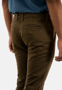 Pantalones de algodón marrones de corte slim, con un bolsillo trasero con cremallera y una etiqueta de marca cerca de la cinturilla. Textura suave.