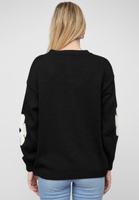Pull noir en maille surdimensionné avec des manches longues, mettant en avant des motifs de fleurs blanches sur les deux bras et une finition texturée.