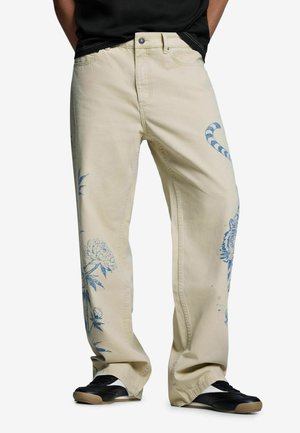 Pantalon beige à jambes larges avec imprimés floraux et tigre bleus, porté avec des baskets noires et blanches et une chemise noire.