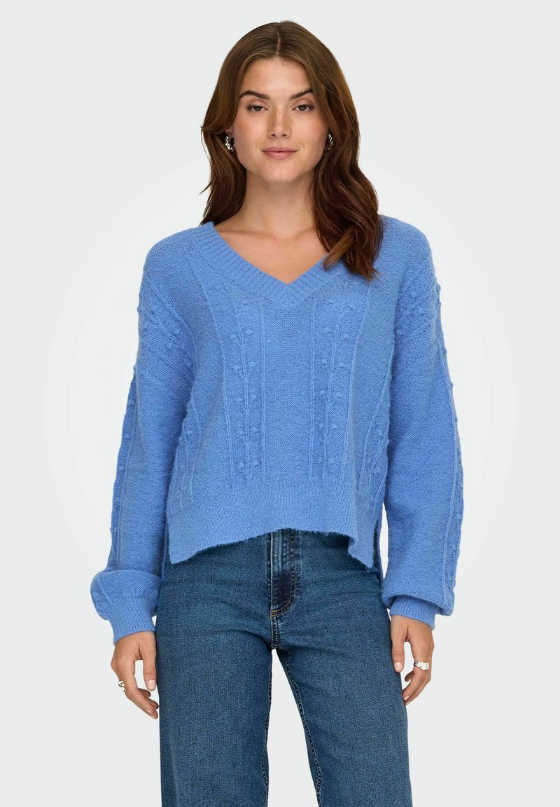 Lichtblauwe gebreide trui met een V-hals, verlaagde schouders en gek textureerde ribbel details. Gestyled met donkere spijkerbroeken.