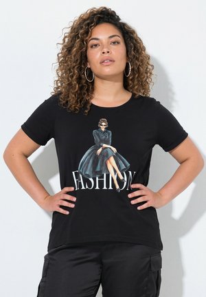 Vrouw met krullend haar en oorringen draagt een zwart T-shirt met een illustratie van een zittende vrouw in een zwarte jurk en het woord "FASHION."