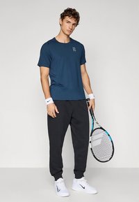 Męski strój sportowy składający się z granatowej koszulki z krótkim rękawem, czarnych spodni dresowych i białych butów, trzymający rakietę tenisową.