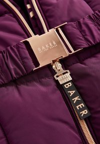 La veste matelassée violette présente une fermeture éclair et une boucle en or brillant avec un logo gravé. Texture lisse avec des détails froncés à la taille.