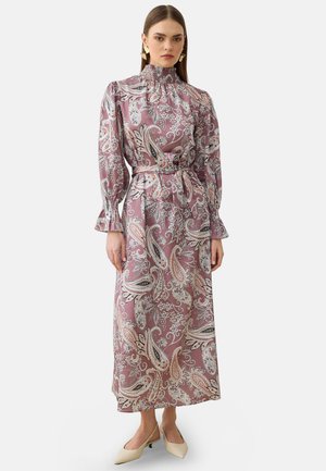 Kvinde iført en langærmet, højhalset pink paisley-kjole med bælte, kombineret med beige lave hælede slingback-sko.