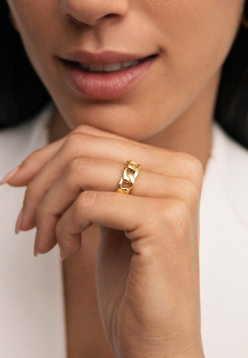 Anello in catena dorata esposto su una mano con finitura naturale delle unghie. La superficie liscia e i collegamenti che si intrecciano creano un interesse visivo.