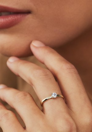 Gouden ring met een gladde band en een ronde, duidelijke edelsteen in het midden. De hand raakt voorzichtig het gezicht aan, waarbij de ring wordt getoond.