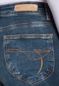 Jeans de mezclilla con un lavado azul oscuro, con costuras naranjas en contraste y un diseño bordado único cerca del bolsillo trasero.