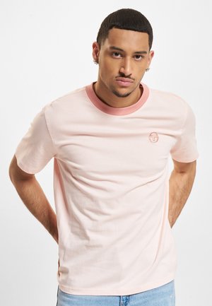 Mann iført lys rosa t-skjorte med mørkere rosa krage og lite logo, poserer med hendene bak ryggen mot hvit bakgrunn.