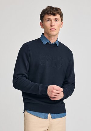 Sudadera azul marino con "POLO CLUB" grabado en el frente, superpuesta a una camisa de mezclilla azul clara, combinada con pantalones beige.