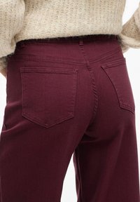 I pantaloni bordeaux presentano un tessuto liscio e strutturato con due tasche posteriori e una vita aderente, parzialmente coperta da un maglione in maglia beige.