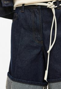 Tamsiai mėlyni džinsiniai šortai su apvyniotais šviesiai pilkais kraštais, baltas raištis diržas, surištas juosmens srityje, ir viršuje matomas džinsinis švarkas.