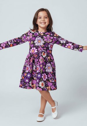 Uśmiechnięta młoda dziewczyna z wyciągniętymi ramionami, ubrana w fioletową sukienkę w kwiaty i białe buty, stojąca na jednej skrzyżowanej nodze na jednolitym tle.