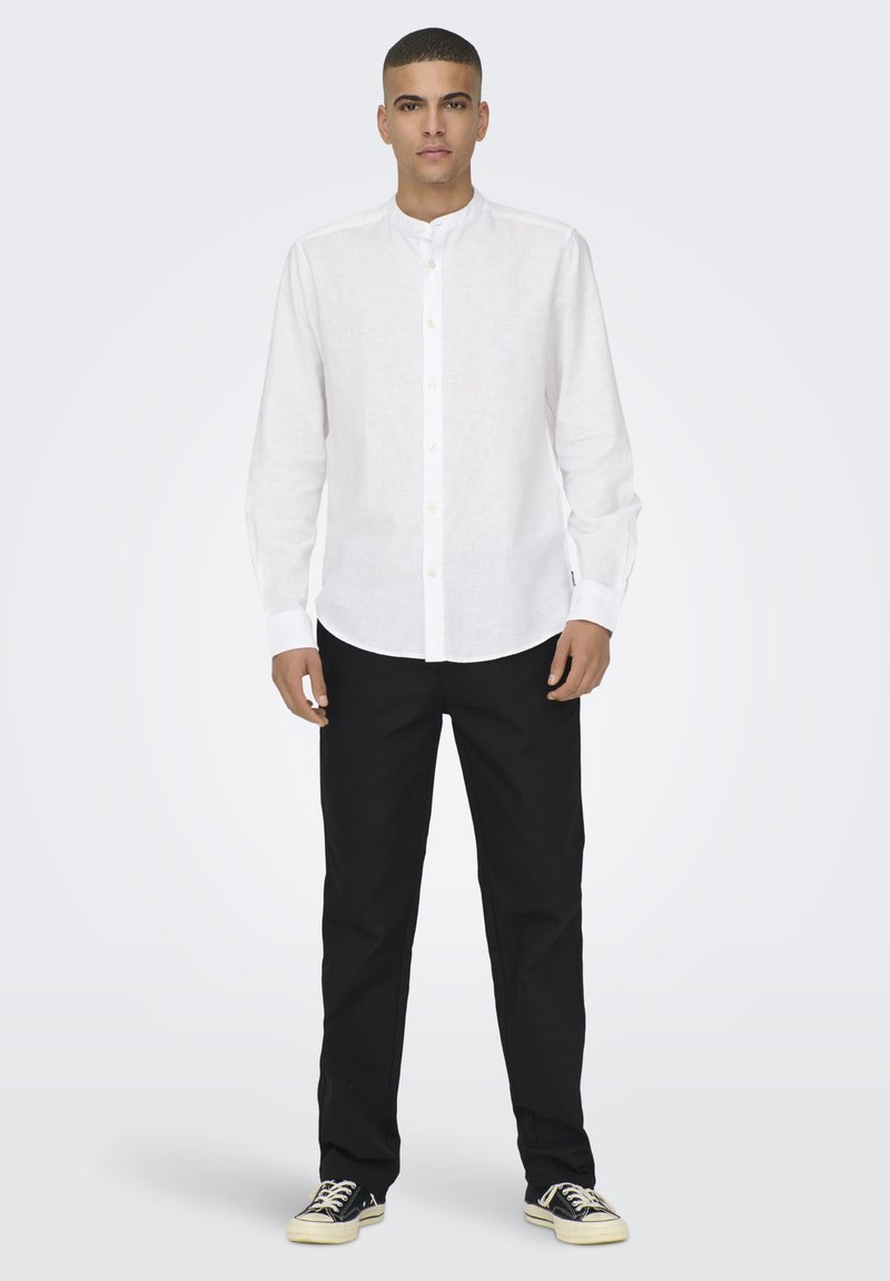 Camicia bianca in lino a maniche lunghe con colletto alla coreana, chiusura con bottoni, e pantaloni neri. Il modello indossa scarpe da ginnastica casual su uno sfondo neutro.