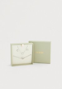 Σετ ασημένια κοσμήματα σε ένα ανοιχτό πράσινο κουτί. Περιλαμβάνει μενταγιόν σε σχήμα καρδιάς και ταιριαστά σκουλαρίκια, όλα με λαμπερό φινίρισμα.