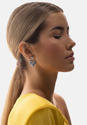 Kobieta z gładkim blond kucykiem, nosząca duże srebrne kolczyki w kształcie serca i żółtą bluzkę z odkrytymi ramionami, o zamkniętych oczach i spokojnym wyrazie twarzy.
