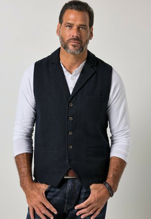 Man with short beard wearing dark vest over white long-sleeve shirt and dark jeans stands with hands near pockets against plain background.