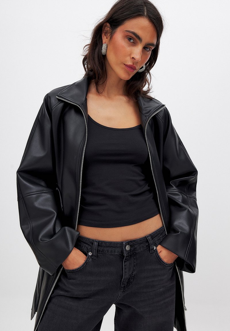 Črna jakna iz umetnega usnja z zadrgo, oversize kroj in širokimi rokavi, nošena čez črno majico brez rokavov ter v kombinaciji s temno modrimi jeans hlačami.
