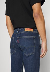 Tummansinisiä farkkuja klassisella leikkauksella. Ominaisuuksiin kuuluu takataskut, keltainen ommel ja nahkapaikka vyötäröllä.