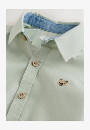 Šviesiai žalia marškinėliai su džinsiniu apykakle ir mediniais sagomis. Ant krūtinės yra maža ruda meškiuko siuvinėjimas. Švelni tekstūra.